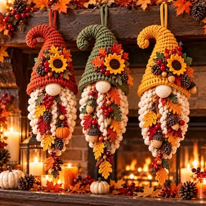 May include: Three hanging crochet gnome ornaments featuring pointed hats in red, green, and yellow. Each gnome has a cream-colored textured beard adorned with crocheted sunflowers, autumn leaves, pinecones, berries, acorns, and small pumpkins.