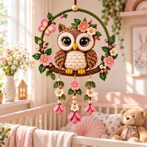 Op de afbeelding: Een gehaakte uil wandhanger met een bruine en crèmekleurige uil in een groene cirkelvormige ring. De ring is versierd met roze en witte gehaakte bloemen, groene bladeren en een roze vlinder. Drie bungelende strengen zijn voorzien van bloemen.