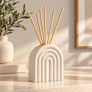 May include: A matte off-white ceramic reed diffuser vessel shaped like a rounded arch with a series of concentric raised ridges. Five thin, light-colored wooden reeds are inserted into the top opening of the structure, extending upwards.