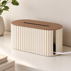 Op de afbeelding: Een ovale kabelmanagementbox met een crèmekleurige, verticaal geribbelde buitenkant en een glad, bruin deksel. De box heeft een smalle rechthoekige gleuf aan de bovenkant en een verticale opening aan de zijkant voor witte elektrische snoeren.