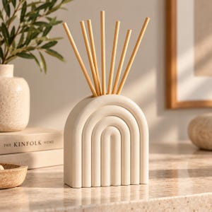 May include: A matte off-white ceramic reed diffuser vessel shaped like a rounded arch with concentric embossed lines. Seven light-colored wooden reeds are inserted into the top opening of the structure, extending upwards in a fan-like arrangement.