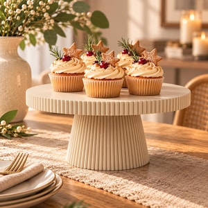 Puede incluir: Un soporte para pasteles de cerámica color crema con base estriada sostiene tres cupcakes decorados con glaseado blanco, bayas rojas, ramitas verdes y galletas en forma de estrella sobre una superficie circular.
