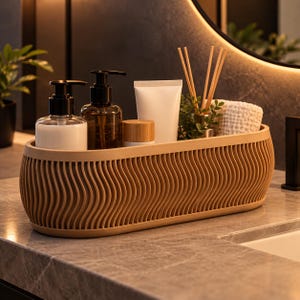 Pode incluir: Uma bandeja de armazenamento oval na cor bege com uma textura externa de ondas verticais. A bandeja contém dois frascos com dispensador, um tubo branco, um pequeno pote com tampa de madeira, uma planta verde, varetas difusoras e um pano branco.