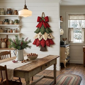 Puede incluir: Una decoraci&oacute;n de pared con forma de &aacute;rbol de Navidad hecha de p&eacute;talos de tela superpuestos en colores rojo, verde y crema, rematada con un gran lazo rojo. El &aacute;rbol presenta detalles bordados de hojas y bayas, un tronco de tela marr&oacute;n y cuerda.