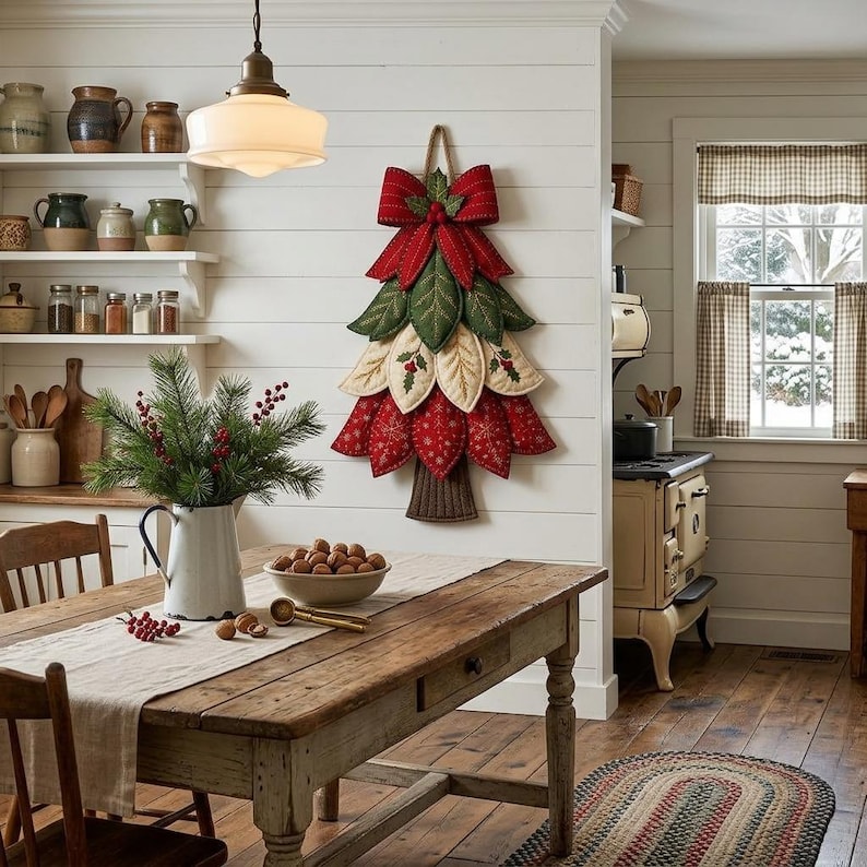 Puede incluir: Una decoraci&oacute;n de pared con forma de &aacute;rbol de Navidad hecha de p&eacute;talos de tela superpuestos en colores rojo, verde y crema, rematada con un gran lazo rojo. El &aacute;rbol presenta detalles bordados de hojas y bayas, un tronco de tela marr&oacute;n y cuerda.