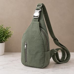 Puede incluir: Una mochila bandolera de lona color verde oliva con forma de lágrima, que cuenta con un bolsillo frontal vertical con cremallera, un compartimento principal con cremallera plateada y una correa ajustable con hebilla metálica.