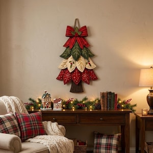 Puede incluir: Una decoraci&oacute;n navide&ntilde;a de pared con forma de &aacute;rbol hecha de p&eacute;talos de tela superpuestos en tonos rojo, verde y crema. El &aacute;rbol presenta motivos bordados de copos de nieve y acebo, rematado con un gran lazo de tela roja y un lazo de yute.