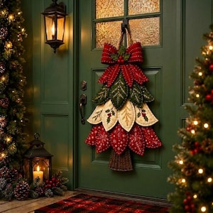 Puede incluir: Una decoraci&oacute;n de pared con forma de &aacute;rbol de Navidad hecha de p&eacute;talos de tela superpuestos en colores rojo, verde oscuro y crema. La pieza presenta un gran lazo rojo en la parte superior, patrones de hojas bordados y un tronco de tela marr&oacute;n.