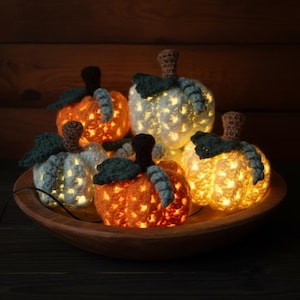 May include: A collection of five crocheted pumpkins in orange and white, each featuring a brown stem and a dark green leaf. These decorative pumpkins are illuminated from within by warm glowing lights and are arranged together in a wooden bowl.