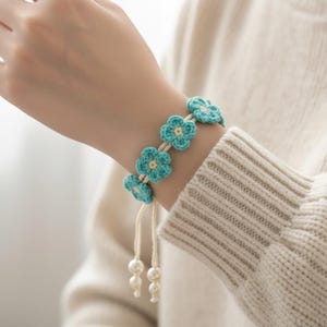 Puede incluir: Una pulsera de ganchillo hecha a mano que presenta una serie de pequeñas flores de color azul claro con centros de color crema. La pulsera está conectada por un cordón fino de color crema e incluye dos extremos colgantes con perlas blancas.