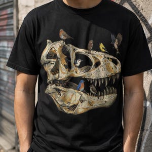 May include: A black short-sleeved t-shirt featuring a large graphic print of a beige dinosaur skull. The skull is decorated with several small, colourful birds perched on and inside the bone structure, including a blue bird and a yellow bird.