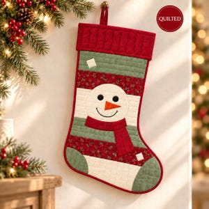 May include: A quilted Christmas stocking featuring a snowman face with a carrot nose, black eyes, and a smile. The stocking has horizontal stripes in red, green, and cream, with a red cuff at the top and a small red circle label reading QUILTED.