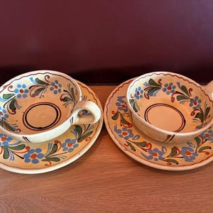 Op de afbeelding: Twee keramische theekopjes met bijpassende schoteltjes op een crèmekleurige basis met handgeschilderde bloemmotieven in blauw, groen en oranje. Elk kopje heeft een enkel zijhandvat en een cirkelvormig ringontwerp op de bodem van de binnenzijde.