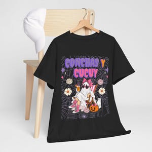 May include: A black t-shirt featuring a graphic print with the text CONCHAS Y CUCUY. The design shows a ghost wearing a pink bow and heart-shaped sunglasses, sitting in a chair with a drink, surrounded by concha bread, flowers, and a pumpkin.