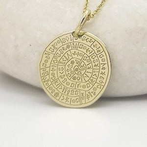 May include: A gold-toned circular pendant necklace featuring a detailed engraved spiral pattern of ancient symbols. The round charm is suspended from a delicate matching gold-toned chain, showcasing a polished metallic finish and intricate etched markings.