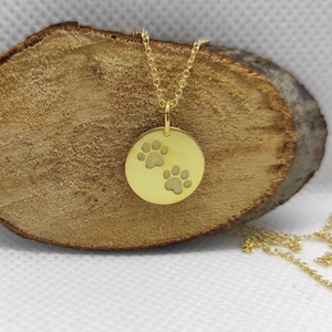 May include: A gold-toned circular pendant necklace featuring two engraved paw prints on the front surface. The pendant is suspended from a delicate gold-toned chain, showcasing a polished metallic finish and a simple, clean design for everyday wear.