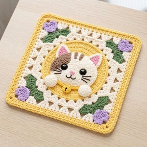 May include: A square crochet coaster featuring a central yellow circle with a white and brown cat face. The cat has black eyes, a pink nose, and a yellow collar with a bell. The border is white lace with green leaves and purple flowers.