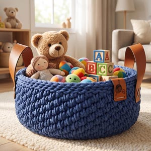 May include: A round, deep blue chunky knit storage basket with two tan leather handles. The basket is filled with a brown teddy bear, a small doll, several colourful round balls, and wooden alphabet blocks featuring the letters A, B, and C.
