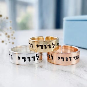 May include: Three open-ended metal rings in silver, gold, and rose gold tones. Each band features a flat surface engraved with black Hebrew text that reads Ani L'dodi V'dodi Li. The rings have a polished finish and a simple, adjustable band design.