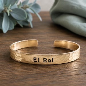 May include: A gold-coloured cuff bracelet with a hammered texture. The bracelet has the words "El Roi" engraved in black. The open-ended design allows for easy wear. The bracelet is displayed on a wooden surface.