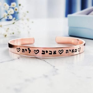 May include: A rose gold cuff bracelet featuring a polished metallic finish and an open design. The exterior surface is engraved with black Hebrew text and small heart symbols interspersed between the words, creating a continuous decorative pattern.