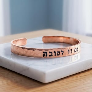 May include: A copper-toned metal cuff bracelet with a hammered texture. The exterior surface features black engraved Hebrew text that reads "גם זו לטובה". The bracelet has an open-ended design, allowing for adjustment to fit the wrist, and displays a metallic finish.