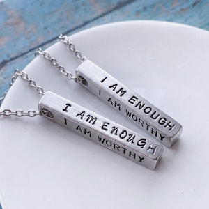 May include: Two silver-toned metal bar necklaces featuring the stamped text I AM ENOUGH and I AM WORTHY on the front faces. Each rectangular pendant is attached to a matching silver-toned metal chain link necklace, displayed together.
