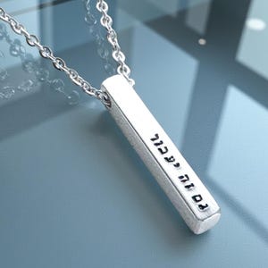 May include: A silver-colored pendant necklace with a rectangular bar pendant. The pendant has engraved text in Hebrew. The chain is a delicate link style. The necklace is displayed on a reflective surface.