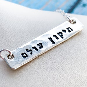 May include: Silver necklace with a hammered bar pendant featuring the Hebrew words "עוצם" and "חזק" in black lettering.