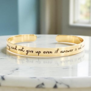 May include: A gold-colored cuff bracelet with the engraved text "pray and not give up even if answer is not". The bracelet is resting on a white marble surface.