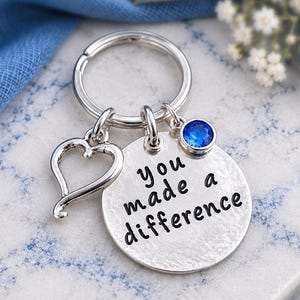 May include: A silver-toned metal keyring featuring a circular disc stamped with the black text 'you made a difference'. The keyring includes a heart-shaped charm and a small, round, bright blue crystal charm attached to a split ring.