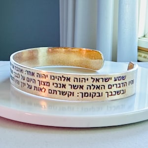 May include: A silver cuff bracelet with Hebrew text. The open-ended bracelet is displayed on a white marble surface. The text is in black and wraps around the outside of the bracelet.