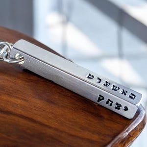 May include: A silver metal keychain with a rectangular bar engraved with Hebrew text. The keychain has a silver ring for attaching keys.