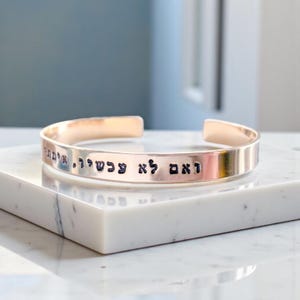 May include: A rose gold cuff bracelet with engraved Hebrew text. The open-ended bracelet is displayed on a white marble surface. The bracelet's polished finish reflects the light, highlighting the engraved inscription.