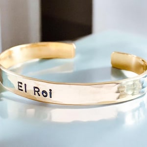 May include: Gold-toned cuff bracelet with the inscription "El Roi" in black lettering. The bracelet has a hammered texture and is open at the ends. The jewelry is displayed on a light blue surface.