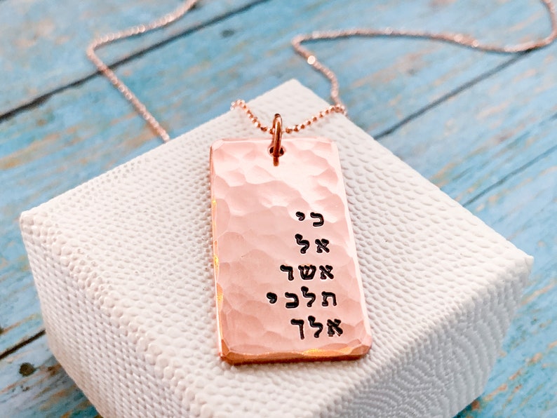 May include: A rose gold necklace with a hammered rectangular pendant featuring Hebrew text. The pendant is engraved with the words "כי אל אשך" which translates to "For God is my light".