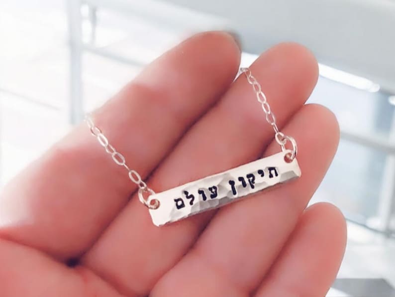 May include: A silver necklace with a hammered bar pendant inscribed with the Hebrew words "עדיף עדיף" (pronounced "adif adif").