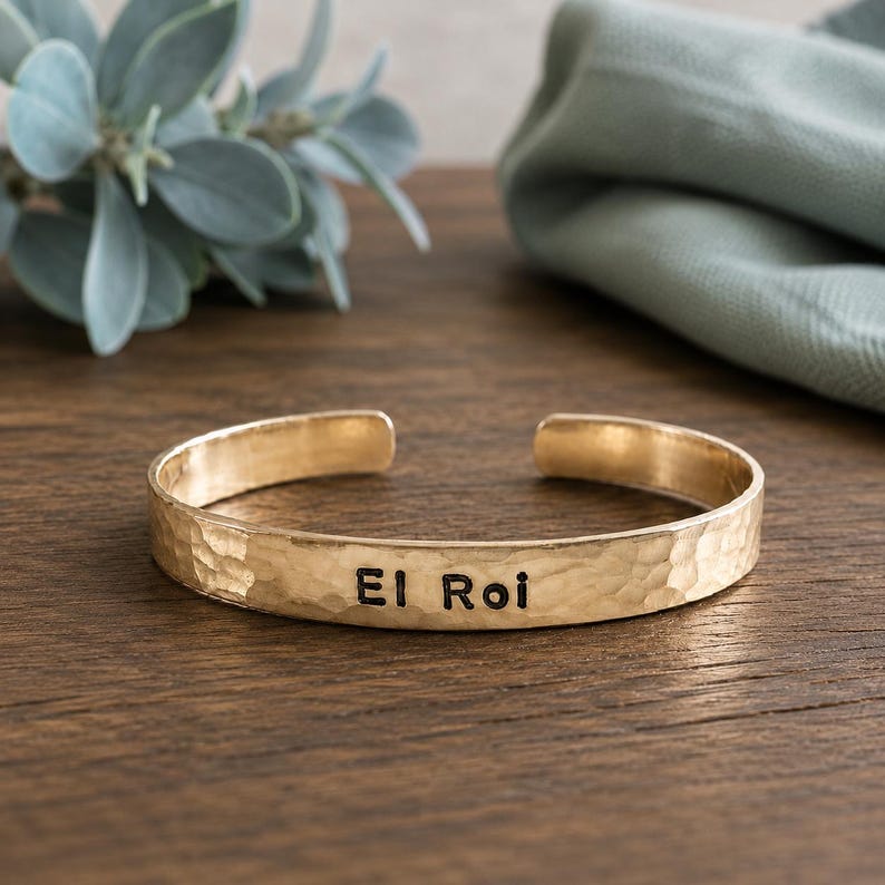 May include: A gold-coloured cuff bracelet with a hammered texture. The bracelet has the words "El Roi" engraved in black. The open-ended design allows for easy wear. The bracelet is displayed on a wooden surface.