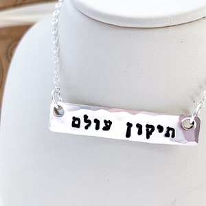 May include: A silver necklace with a hammered bar pendant that reads "הַקָּדוֹשׁ עָלָיִם" in Hebrew.