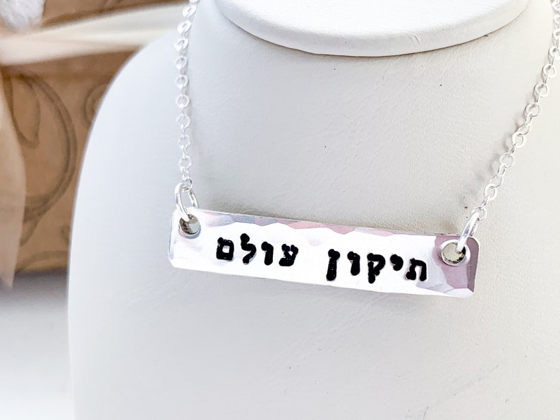 May include: A silver necklace with a hammered bar pendant that reads "הַקָּדוֹשׁ עָלָיִם" in Hebrew.