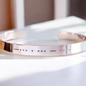 May include: A silver bangle bracelet with a morse code message engraved on the front. The message is "-" "."" "."" "-" "."" "."" "-" "."" "."