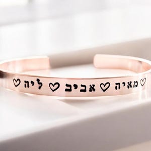 May include: A rose gold bangle bracelet with the Hebrew words "אבא" (father), "אחי" (brother), and "לי" (to me) engraved on it, separated by heart symbols.