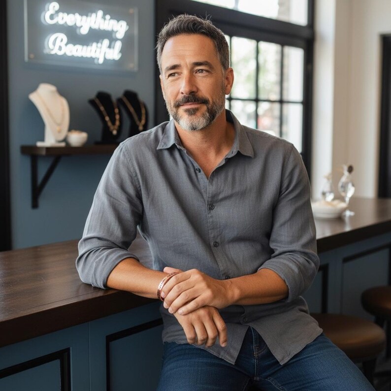 May include: A man in a grey button-down shirt and blue jeans leans on a counter. He wears a copper-coloured bracelet. The background includes jewellery displays and a neon sign that reads "Everything Beautiful."