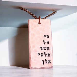 May include: A pink rectangular tag with Hebrew text hanging from a brown chain. The tag has a hammered metal texture. The text reads "כי אל אשך הכה כל"