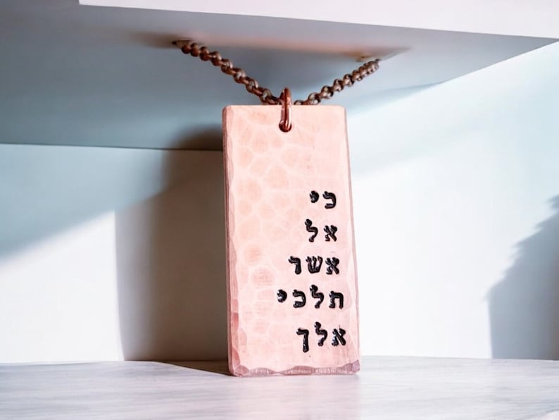May include: A pink rectangular tag with Hebrew text hanging from a brown chain. The tag has a hammered metal texture. The text reads "כי אל אשך הכה כל"