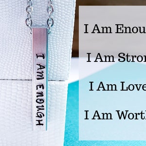 A silver, 3D, rectangle pendant on a display box. The front side of the pendant is hand stamped with the words I am enough in a cute font. The photo tag reads I am strong, I am loved, and I am worthy. It hangs from a silver regular chain.