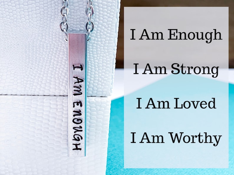 A silver, 3D, rectangle pendant on a display box. The front side of the pendant is hand stamped with the words I am enough in a cute font. The photo tag reads I am strong, I am loved, and I am worthy. It hangs from a silver regular chain.