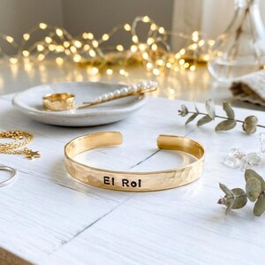May include: A gold-coloured hammered cuff bracelet inscribed with "El Roi". The bracelet is displayed with other gold jewellery, including a ring, a necklace, and a hair clip with pearls. The items are on a white surface.
