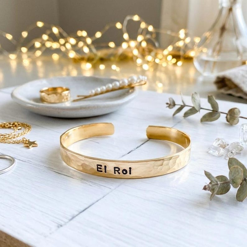 May include: A gold-coloured hammered cuff bracelet inscribed with "El Roi". The bracelet is displayed with other gold jewellery, including a ring, a necklace, and a hair clip with pearls. The items are on a white surface.