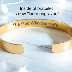 May include: Gold-coloured bracelet with the engraved text "The God Who Sees Me." The inside of the bracelet is laser engraved. The bracelet has a hammered texture and is open-ended.